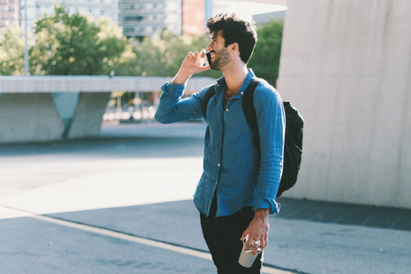 attractive young guy with a backpack wearing in a casual clothes talking to a friend on a smartphone. professional manager  discusses the details of a new project by phone on the way to the office.の写真素材