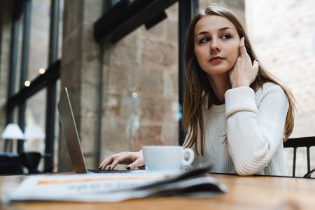 A caucasian female freelancer is looking aside while sitting in a cafe with a portable computer and a cup of coffee on a table. Pensive european girl sitting with electronic devices in a cafe.の写真素材
