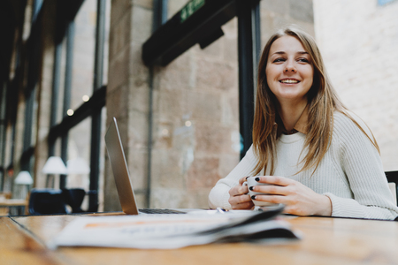 Beautiful caucasian woman dreaming about something while sitting with portable computer in a modern cafe. Young charming female freelancer smiling while thinking new good idea for the work.の写真素材