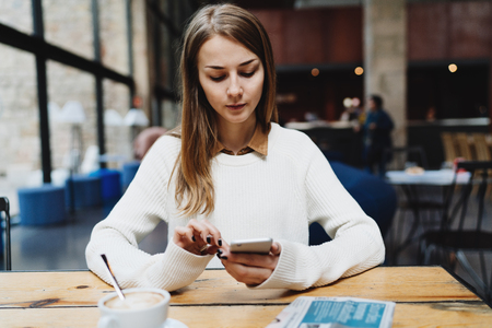 Thoughtful successful business woman reading business emails on a smartphone during coffee break. Hipster girl resting in a coffee shop watching videos online on a display of her mobile phone.の写真素材