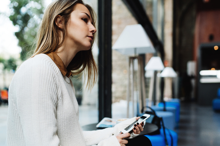 Portrait of charming blonde woman sitting beside a big window of a modern coffee shop with a mobile phone in her hands. Thoughtful student girl holding smartphone while sitting on a blurred backgroundの写真素材
