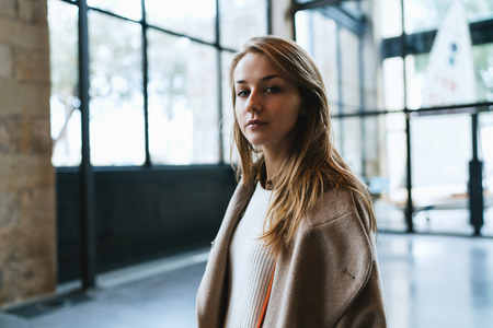 half length portrait of a beautiful blonde hipster girl looking at the camera while standing half turned to the camera on a blurred background. Student girl wearing stylish clothes walking the city.の写真素材