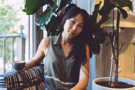 Young asian girl with long dark hair wearing casual clothes is sitting in a cafe while having rest. A restaurant blogger with a cup of tea is sitting on a sofa and looking at the camera.の写真素材