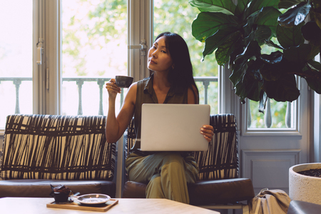 Beautiful asian woman with long dark hair is drinking tea while sitting with a portable computer in a modern coffee shop. Young entrepreneur is waiting for the informal business meetng in a restaurantの写真素材