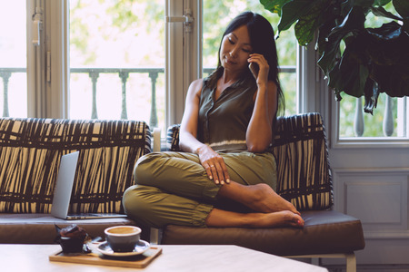 Young college student girl is studying at home on a sunny day. Beautiful asian female with long dark hair is talking by a mobile phone while sitting on a sofa with a laptop on it.の写真素材