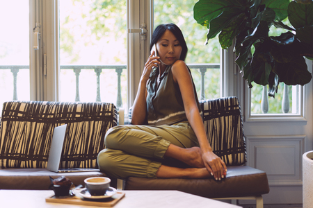 Attractive asian student is talking by mobile phone while sitting in a cafe with the modern interior. Beautiful entrepreneur female wearing casual clothes is making a business call during the break.の写真素材