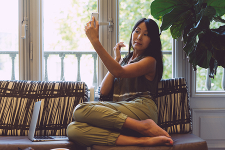 Charming asian woman is using front camera of a smartphone as a mirror to see that her make-up is ok. Young female is holding a mobile phone while sitting with a laptop on a sofa in a modern cafe.の写真素材