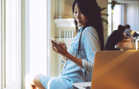 Beautiful asian woman with long dark hair is reading messages on her mobile phone. Young model look woman is chatting by a smartphone while sitting in a modern coffee shop with a portable computer.の写真素材