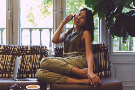 A charming asian travel blogger is talking by mobile phone while sitting in a cafe with the modern interior. Attractive business woman wearing trendy clothes is talking with a business partner.の写真素材