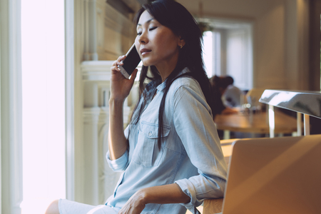 Beautiful asian woman with long dark hair is having a call on her mobile phone. Young model look woman is talking by a smartphone while sitting in a modern coffee shop with a portable computer.の写真素材