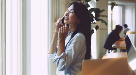 Charming asian woman with long dark hair is talking with friends by her mobile phone. Young business lady is talking by a smartphone while sitting in a modern coffee shop with a portable computer.の写真素材
