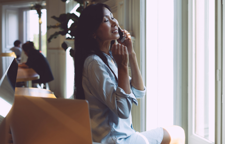 Beautiful asian female with long dark hair is having a call on her mobile phone. Young marketing specialist is talking by a smartphone while sitting in a modern coffee shop with a portable computer.の写真素材