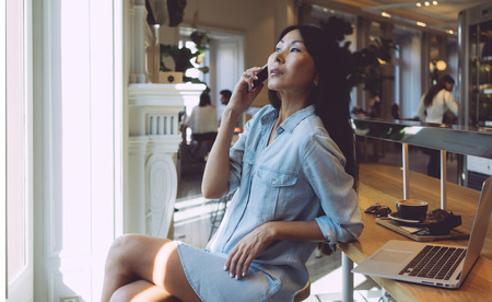 Young entrepreneur is having business conversation on a mobile phone while sitting in a modern coffee shop. Asian woman is shopping online by using a smartphone and a laptop connected to wi-fi.の写真素材