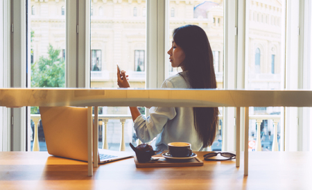Young entrepreneur is reading business emails by a mobile phone while sitting in front of the big window in a modern coffee shop. Asian woman is shopping online by using a smartphone and a laptop.の写真素材