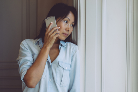 Beautiful asian woman with long dark hair is talking by a smartphone while looking thoughtfully out the window. Hipster girl is chatting with friends by a mobile phone while standing beside the windowの写真素材