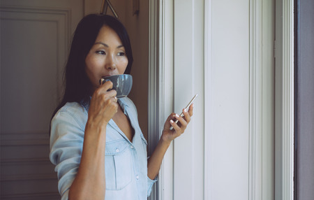 Young entrepreneur is answering business emails by a mobile phone while having break with a cup of tea. Asian woman is holding a smartphone while standing beside the window with a cup of coffee.の写真素材