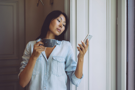 Attractive asian woman is surfing the web by a mobile phone while having break with cup of coffee in modern coffee shop. Modern designer is reading emails on a smartphone connected to wi-fiの写真素材