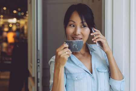 Beautiful asian woman is talking by a mobile phone while having break with cup of coffee in a modern coffee shop. Young entrepreneur is appointing the meeting with the business partners by phone call.の写真素材