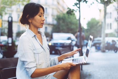 Young asian woman is sitting at city public space with a laptop connected to wi-fi. Entrepreneur female is working on a portable computer while sitting outdoors on a blurred urban background.の写真素材