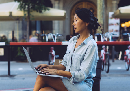 Young asian female is sitting at city public space with a laptop connected to wi-fi. Business woman is working on a portable computer while sitting outdoors on a blurred urban background.の写真素材