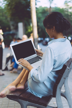 Beautiful asian woman is sitting at city public space with a laptop connected to wi-fi. Young web designer is working on a portable computer while sitting outdoors on a blurred urban background.の写真素材