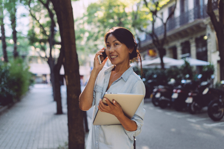 Asian business woman is having business call by a smartphone while standing with a laptop in her hand on a blurred urban background. Beauty blogger is going down the street holding digital devices.の写真素材