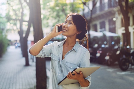 Asian entrepreneur female is having call by a mobile phone while standing with a laptop in her hand on a blurred urban background. Cute student girl is going down the street holding digital devices.の写真素材