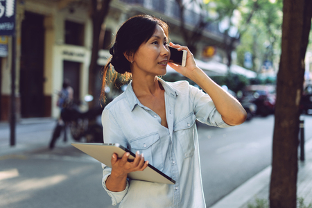 Asian business woman is having business call by a mobile phone while standing with a laptop in her hand on blurred urban background. Cute hipster girl is going down the street holding digital devicesの写真素材