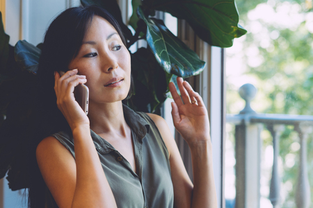 Beautiful asian female is talking on a mobile phone while sitting in the modern coworking space. Young entrepreneur is calling to the clients while having breakfast in a coffee shop.の写真素材
