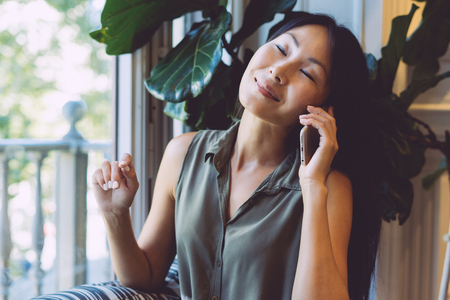 Beautiful asian woman is talking on a mobile phone while sitting in the modern coworking space. Young freelancer girl is calling to the clients while having breakfast in a coffee shop.の写真素材