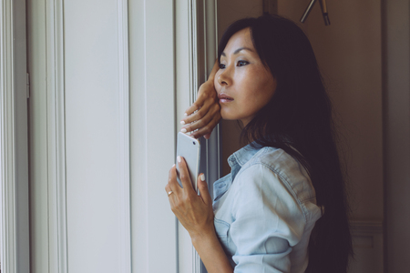A beautiful asian female is holding a smartphone while looking at the big window with a cup of coffee in the morning. Beauty blogger is reading news on a smartphone during the brunch.の写真素材
