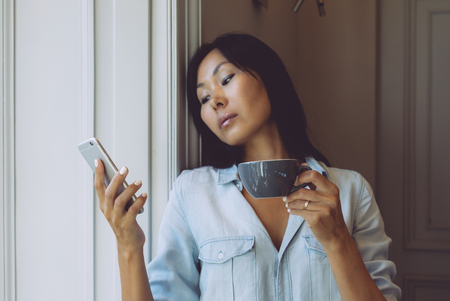A thoughtful asian female is holding a smartphone while standing at the big window with a cup of coffee in the morning. Fashion blogger is reading news on a smartphone during the breakfast.の写真素材