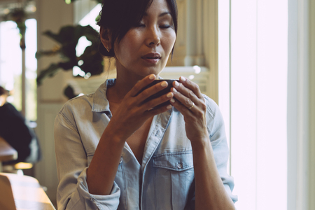 A portrait of a beautiful asian female with long dark hair wearing light blue jean summer dress and sitting in a coffee shop with a cup of tea on a blurred cafe background in the morning.の写真素材