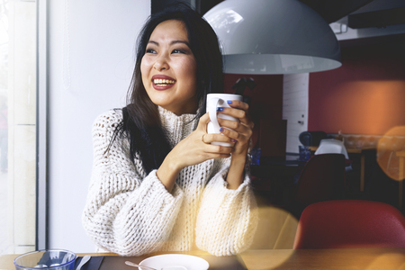 charming girl with a beautiful smile drinking tea sitting in a cozy cafe near the window. happy asian girl smiling and looking out the window cafe. Bokeh light, flareの写真素材