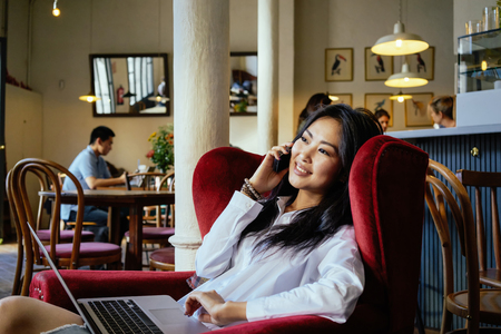 Beautiful Asian business woman at a coffee shop working on a laptop and talking on the smart-phoneの写真素材