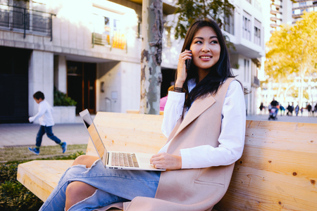 Portrait of a beautiful young smiling asian woman holding on her portable laptop computer while sitting on a wooden bench and talking on the smart-phoneの写真素材