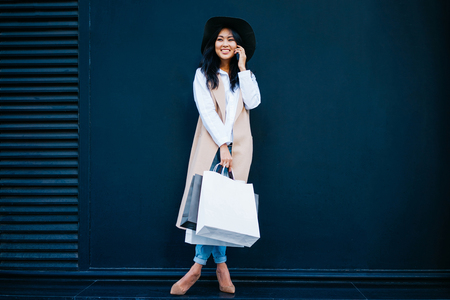 beautiful young Asian businesswoman talking on the phone and holding white blank bags in her hand on the background of black wall with copy space for your text or design.の写真素材