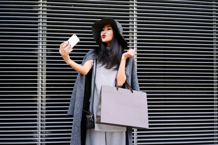 young beautiful stylishly-dressed Asian girl posing and holding a blank paper bag with empty space for your text or logo. Attractive well-dressed woman making selfieの写真素材