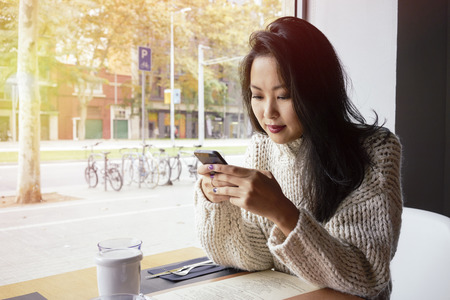 Young Asian Girl drinking coffee / tea and using digital smart-phone in a coffee shop.Flare lightの写真素材