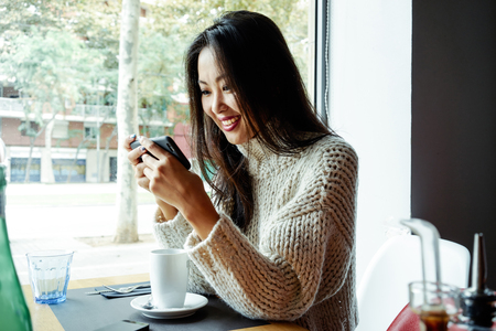 Young Asian Girl drinking coffee / tea and using digital smart-phone in a coffee shopの写真素材