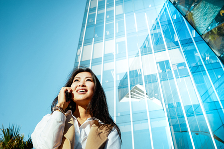 Beautiful Asian business woman making a phone call outside business center.portrait of a attractive girl talking on cell phone on a skyscraper backgroundの写真素材