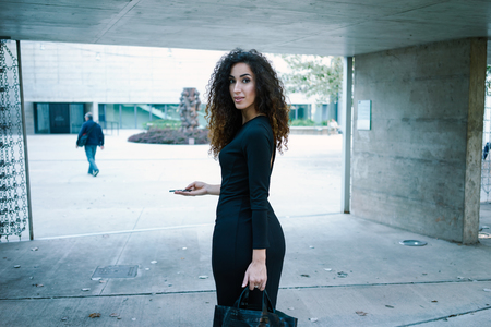 Young attractive business woman goes on the job. young woman with long curly hair in a black fitting dress with gorgeous figure holding a smart-phone against a background of a modern buildingの写真素材