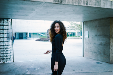 Young attractive business woman goes on the job. gorgeous woman with long curly hair in a black fitting dress holding a smart-phone against a background of a modern buildingの写真素材