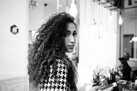 black and white portrait of a young beautiful businesswoman with long curly hair sending text message from her digital smart-phone in coffee-shopの写真素材