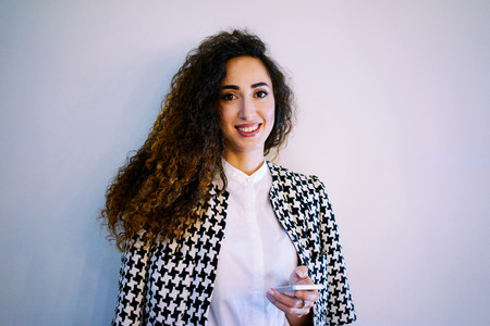 Portrait of a young gorgeous woman sending text message from her digital smart-phone on a white wall backgroundの写真素材