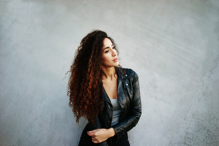 portrait of young beautiful woman with long curly hair wearing a black leather jacket posing on a background of gray concrete wall with copy space area for your text or designの写真素材