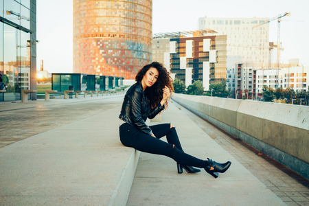 Full length portrait of gorgeous young woman with long curly hair sitting on the stairs against the backdrop of the cityの写真素材