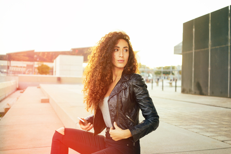 attractive young girl with long curly hair wearing a black leather jacket holding smart-phone on a city buildings background.pretty woman resting outdoors and looking away. flare lightの写真素材