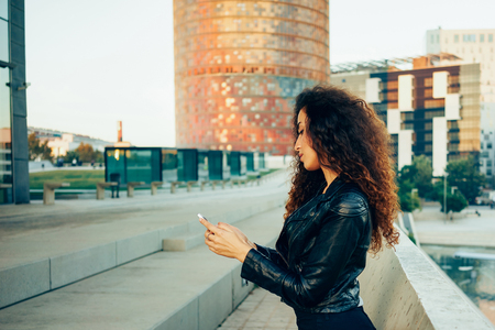 attractive young woman with long curly hair wearing a black leather jacket holding a mobile phone on the background of urban buildings. beautiful young hipster girl using cell telephoneの写真素材