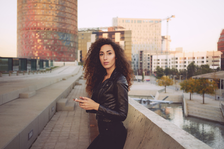 attractive young woman with long curly hair wearing a black leather jacket holding a mobile phone on the background of urban buildings.の写真素材